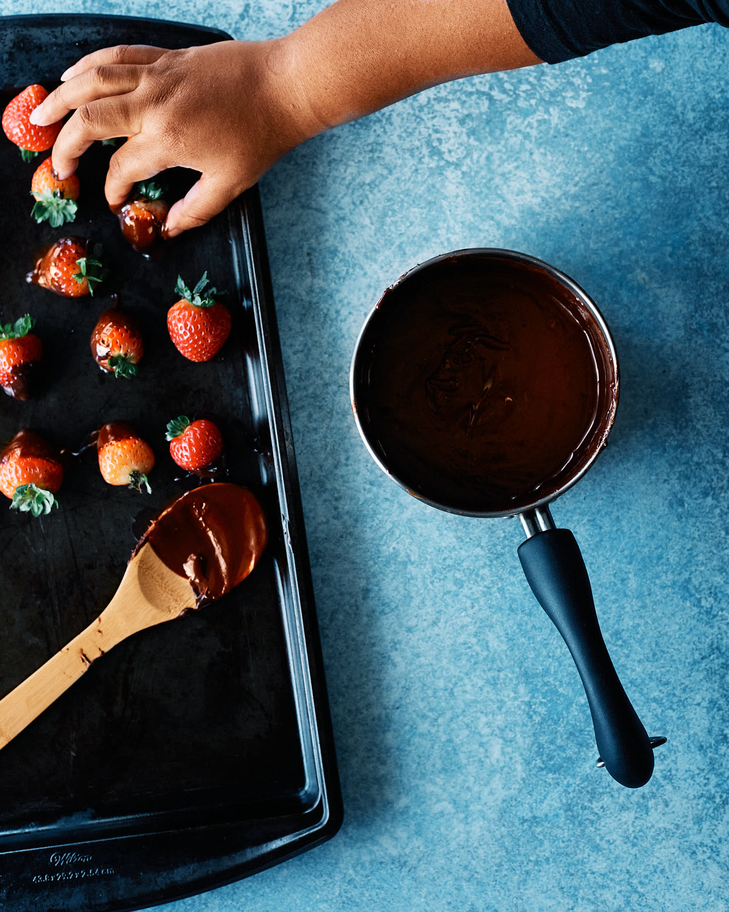 Chocolate covered strawberries on a baking pan and a pot of melted chocolate on a blue background