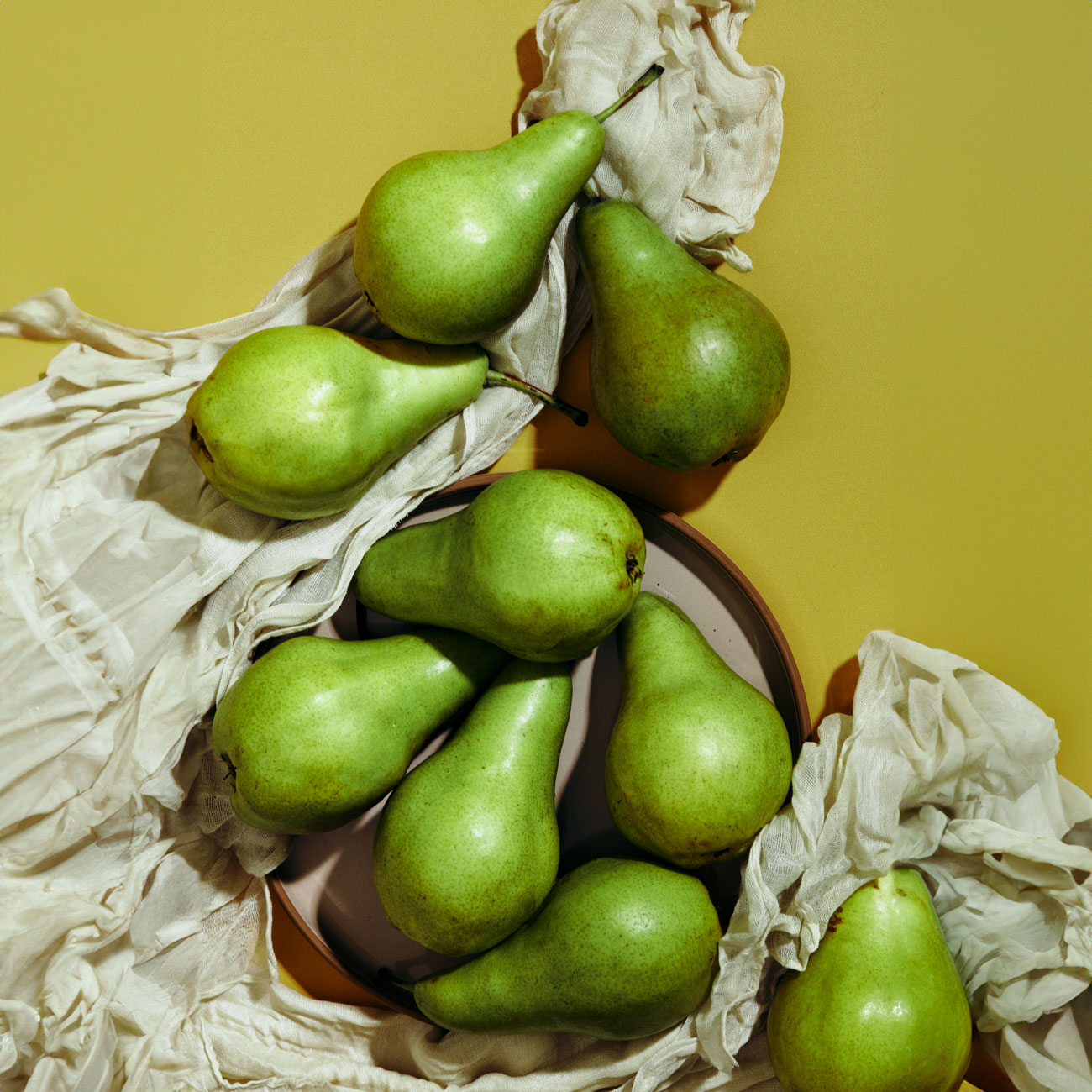 Lush green pears artfully arranged on and around a plate, interspersed with crinkled white cloth, against a mustard yellow backdrop.