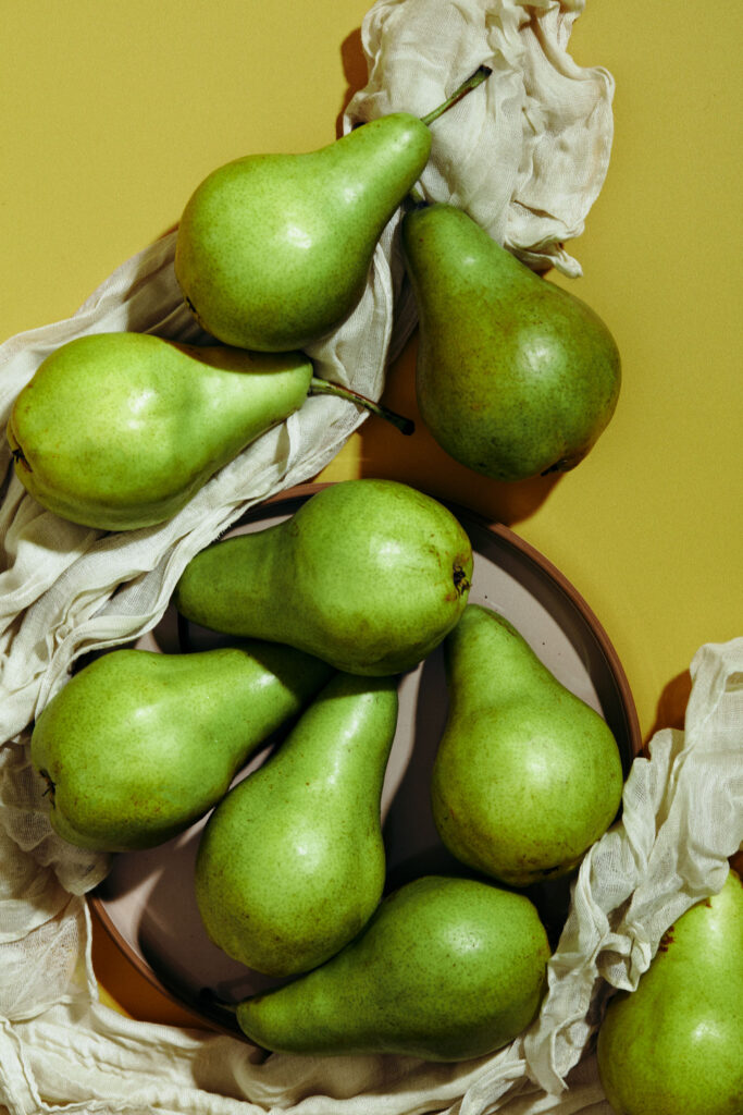 Close-up of fresh green pears arranged amidst draped white cloth on a plate, set against a soft yellow background.