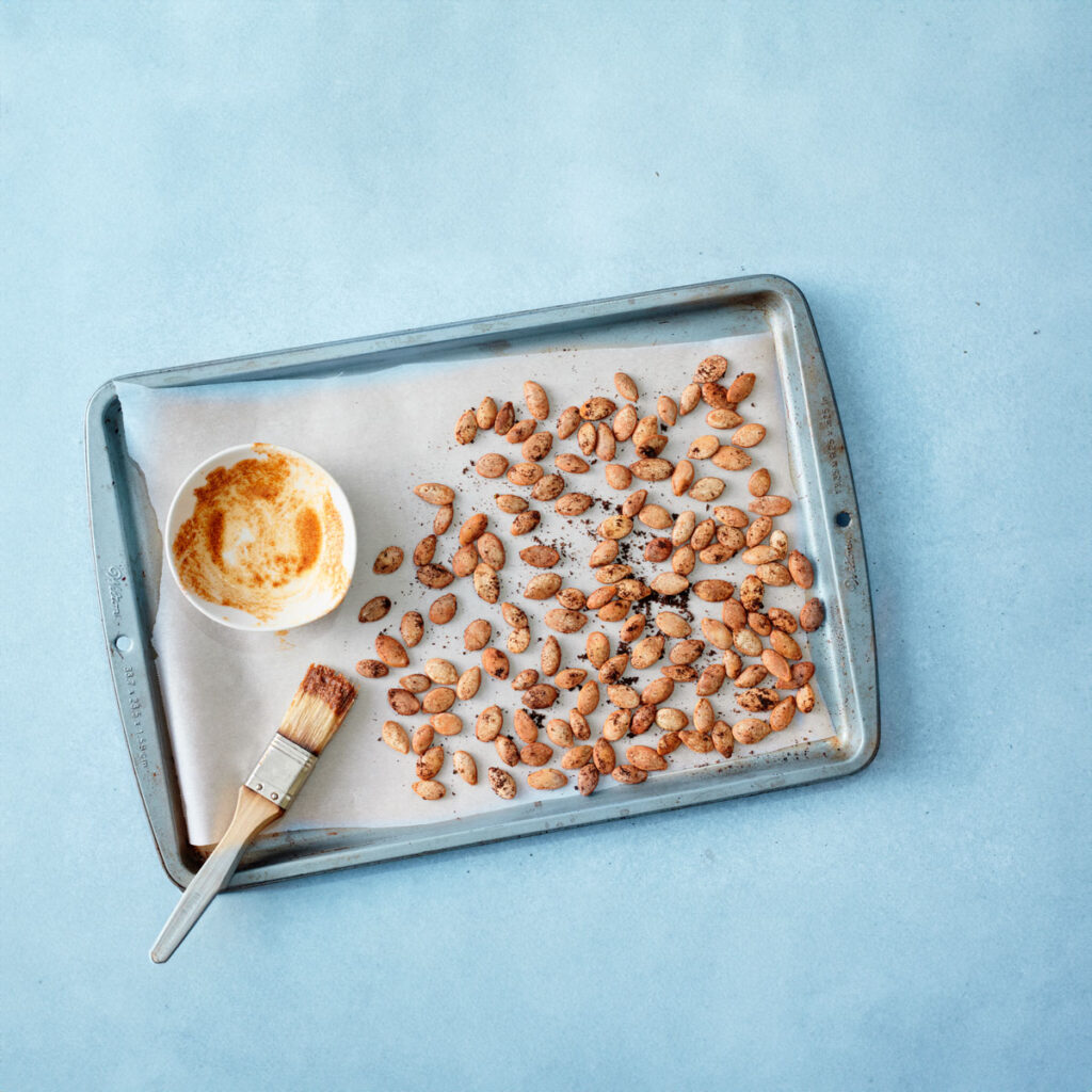 A baking tray with scattered roasted pumpkin seeds, next to an empty bowl with remnants of seasoning and a brush, all set against a light blue background.