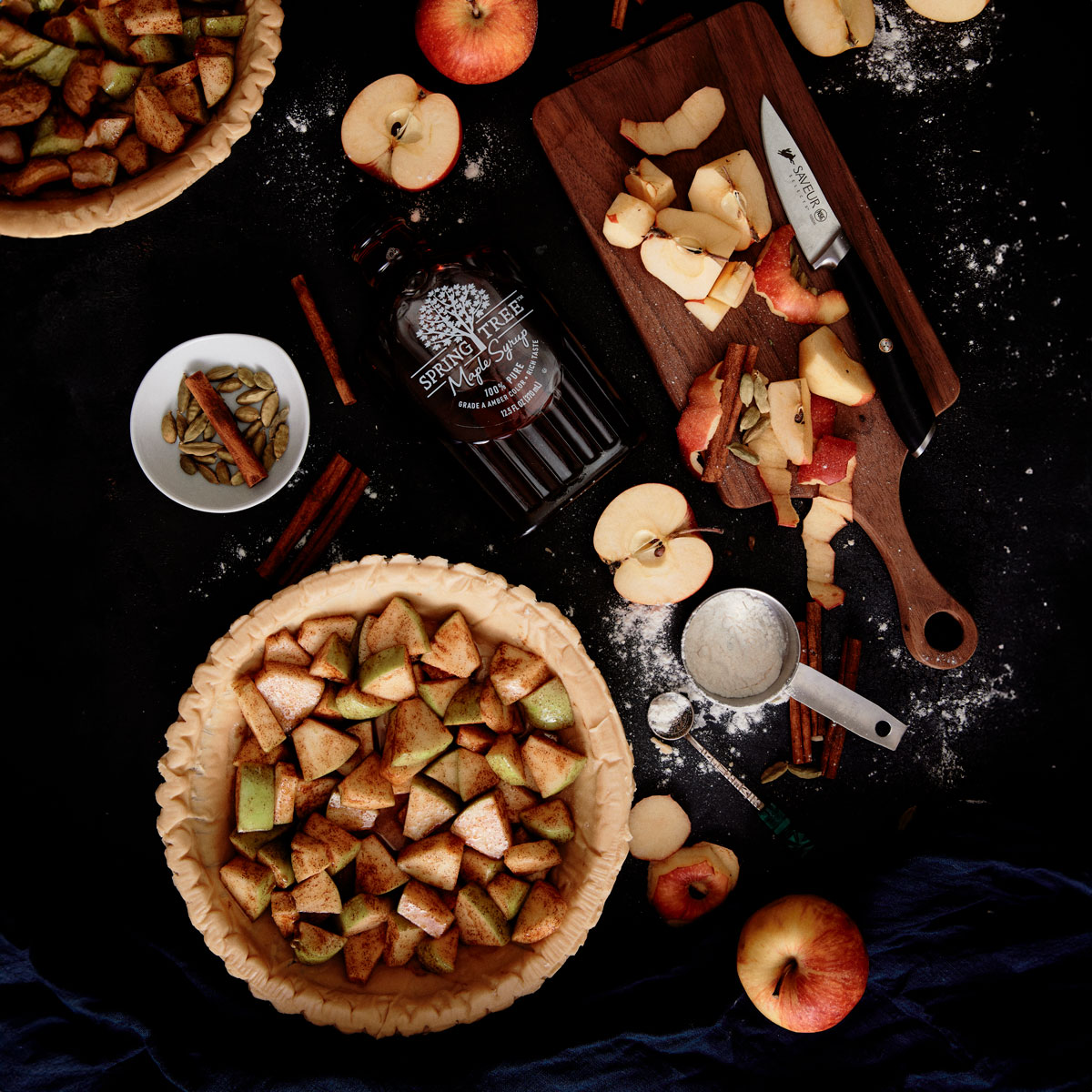 A top-down view of a baking scene featuring an unbaked apple pie filled with diced apples, a cutting board with apple slices and a knife, a bottle of maple syrup, whole and sliced apples, a small bowl with spices, and a scoop of flour, all scattered on a dark surface with hints of flour and a navy cloth.