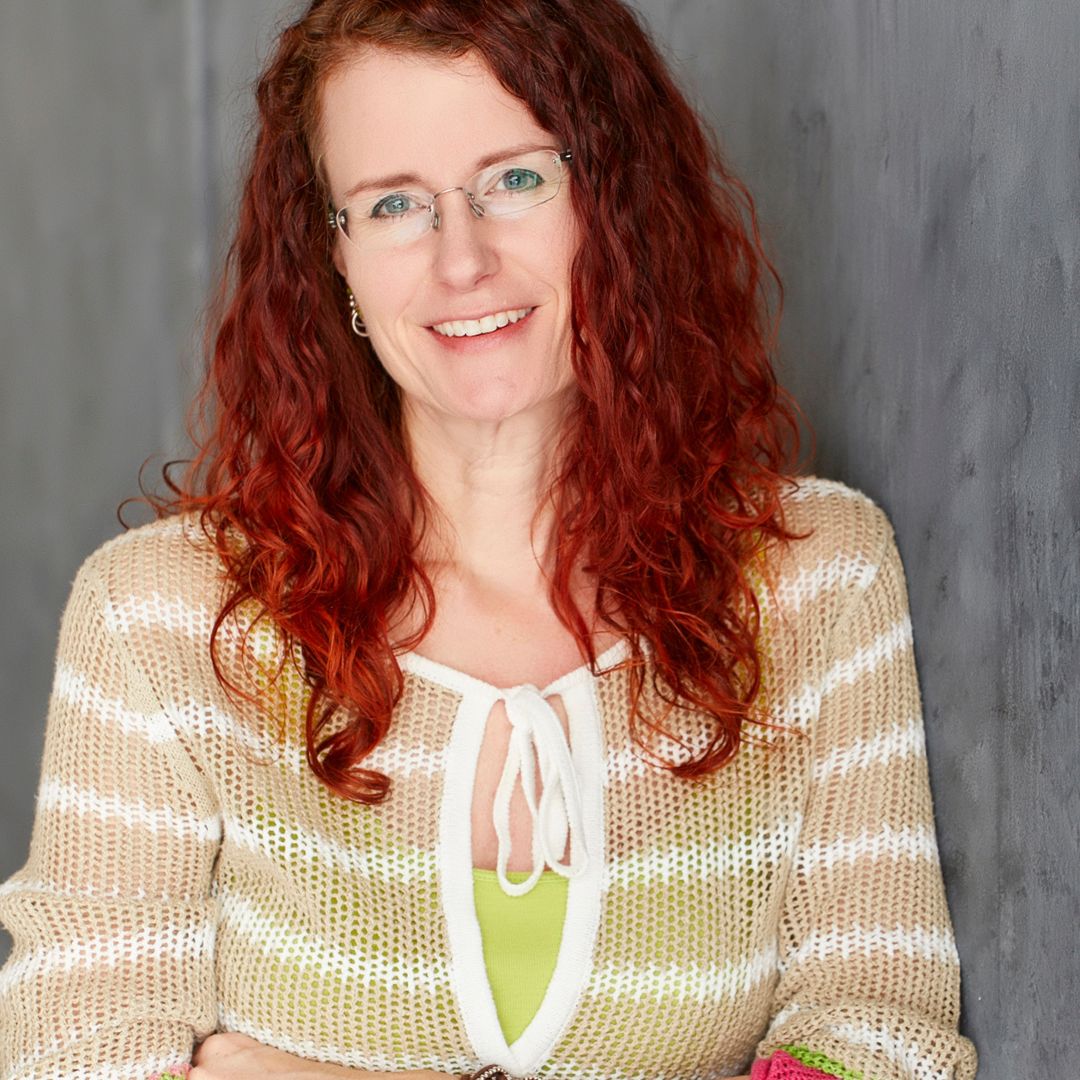Portrait of Christina Peters, smiling with red hair and glasses, wearing a beige striped sweater, against a muted gray background.