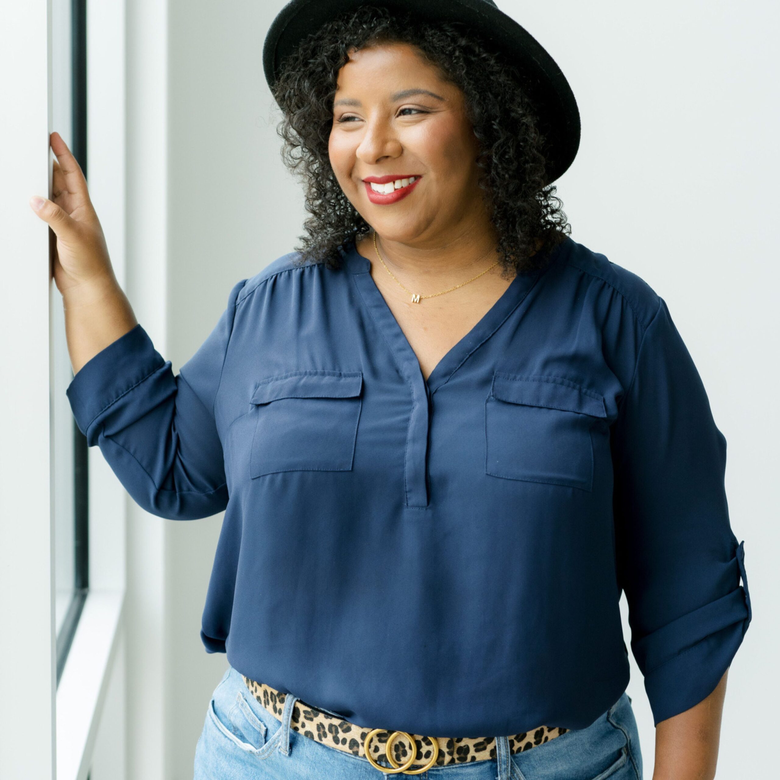 Woman in a blue blouse and black hat smiling by a window.