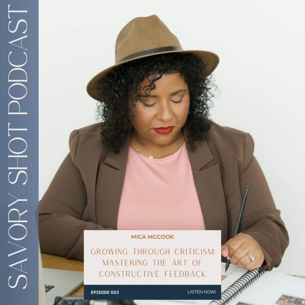 Woman in a brown blazer and hat writing in a notebook at a desk with a laptop and camera.