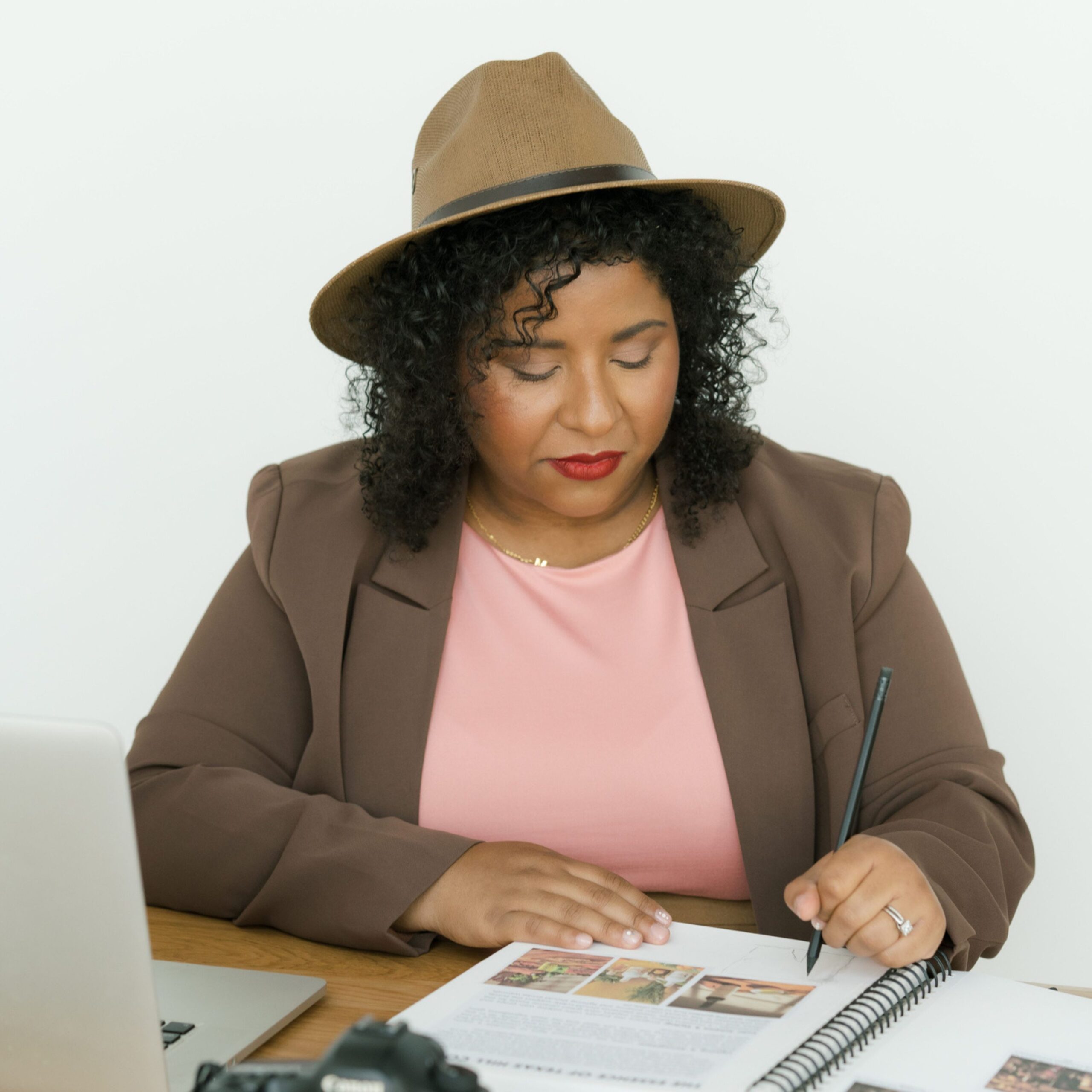 Woman in a brown blazer and hat writing in a notebook at a desk with a laptop and camera.
